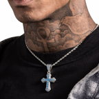 A close-up of a tattooed man wearing a silver chain necklace with a cross pendant featuring light blue inlay, over a black t-shirt and a gray denim jacket.