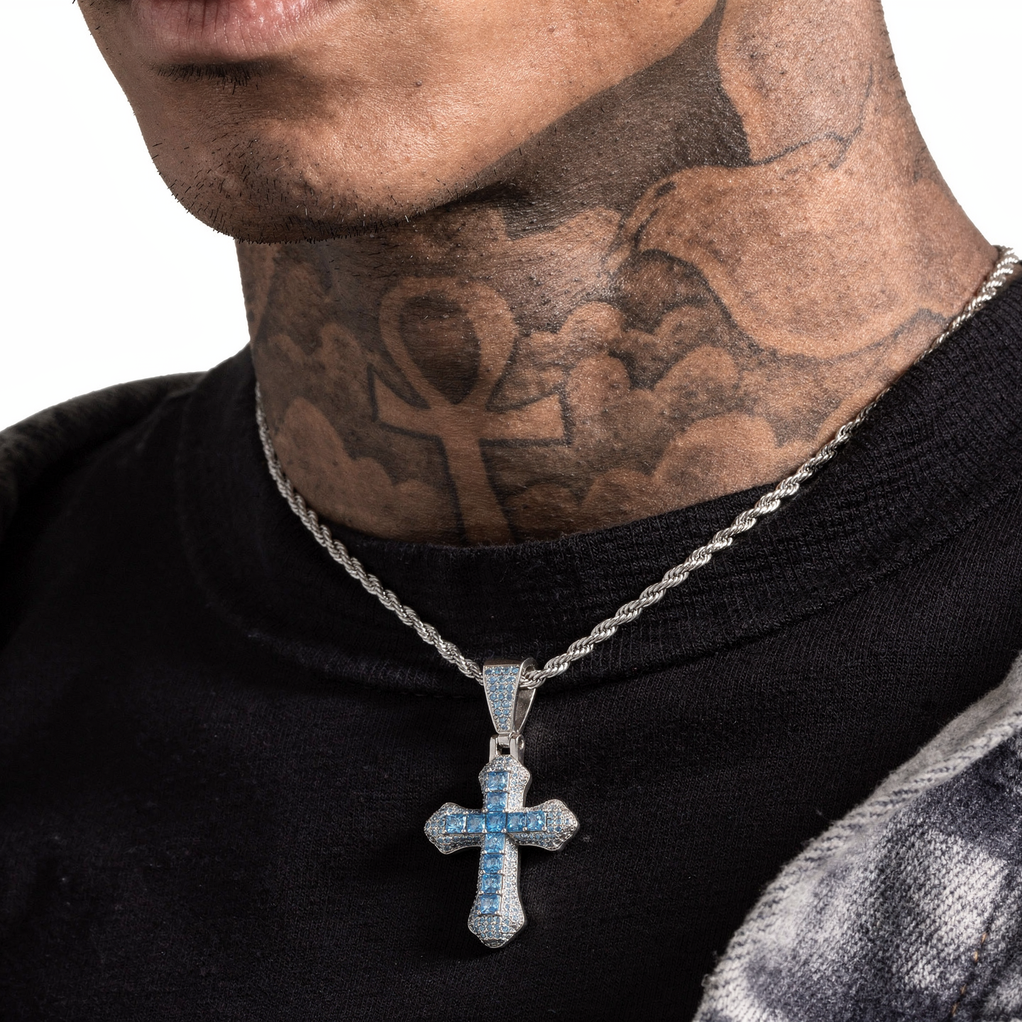 A close-up of a tattooed man wearing a silver chain necklace with a cross pendant featuring light blue inlay, over a black t-shirt and a gray denim jacket.